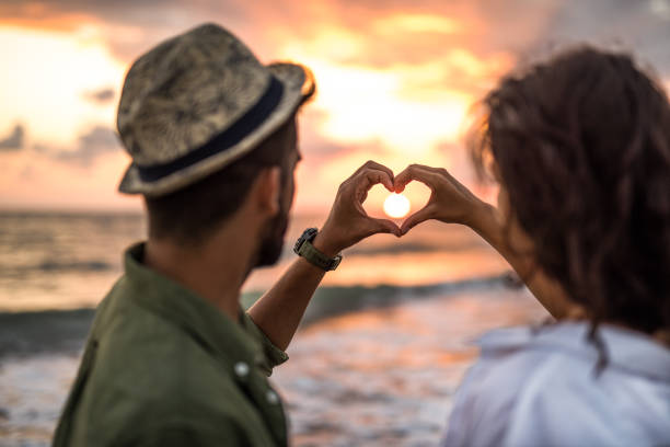 Пара сложила ладони в виде сердца на фоне заходящего над морем солнца